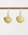 A pair of silver drop earrings with smooth, polished light yellow-green oval stones, hanging from a horizontal brass rod against a light gray background.