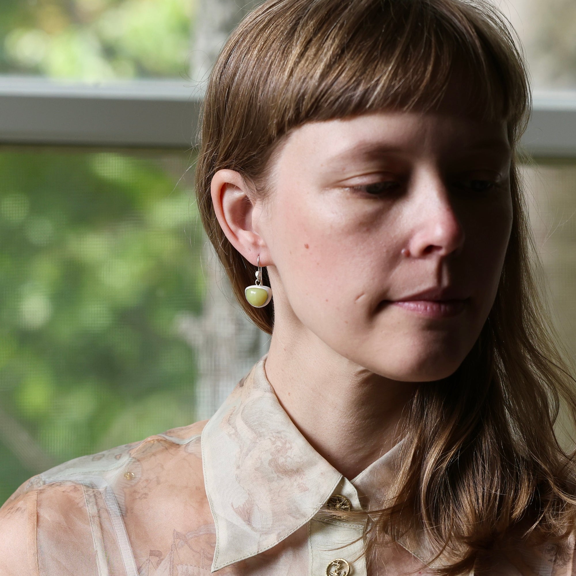 A close-up portrait of a woman wearing a small gold hoop earring with a pale yellow heart-shaped stone dangle, set against a softly blurred window background.