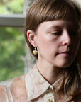 A close-up portrait of a woman wearing a small gold hoop earring with a pale yellow heart-shaped stone dangle, set against a softly blurred window background.