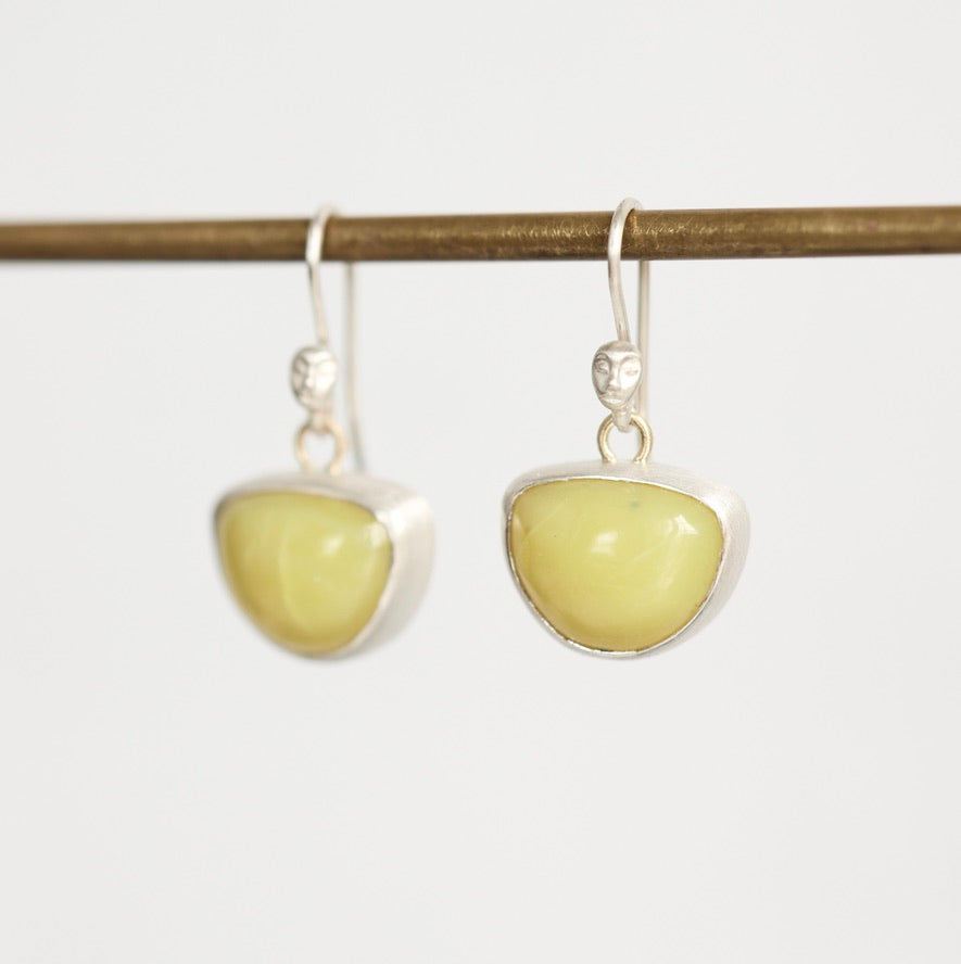 A pair of silver dangle earrings with smooth, polished light yellow-green stone pendants, hanging from a horizontal brass rod against a plain white background.