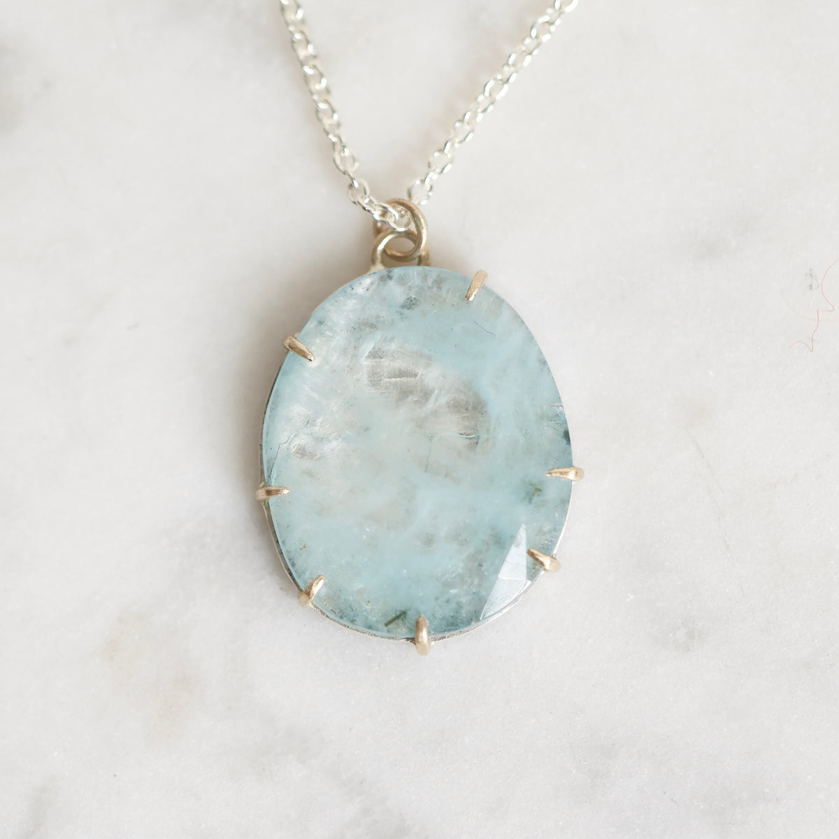 A silver chain necklace with an oval light blue gemstone pendant, held in a gold-toned bezel setting, displayed on a light marble surface.