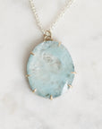 A silver chain necklace with an oval light blue gemstone pendant, held in a gold-toned bezel setting, displayed on a light marble surface.