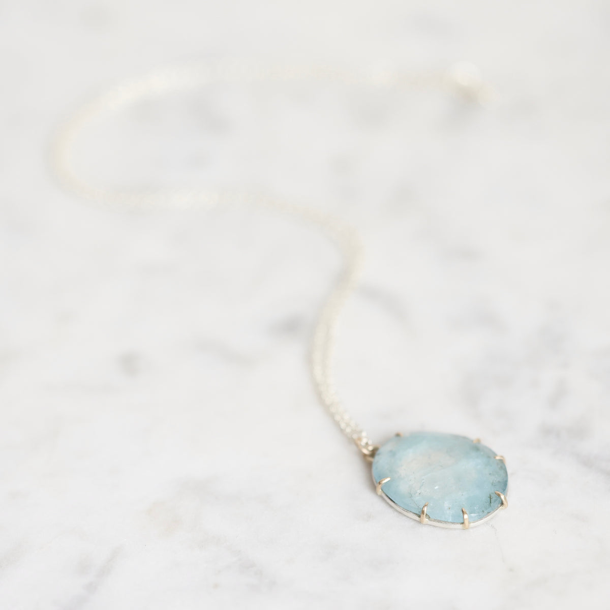 A delicate silver chain necklace with a round, light blue stone pendant set in a gold-toned bezel, resting on a white marble surface.