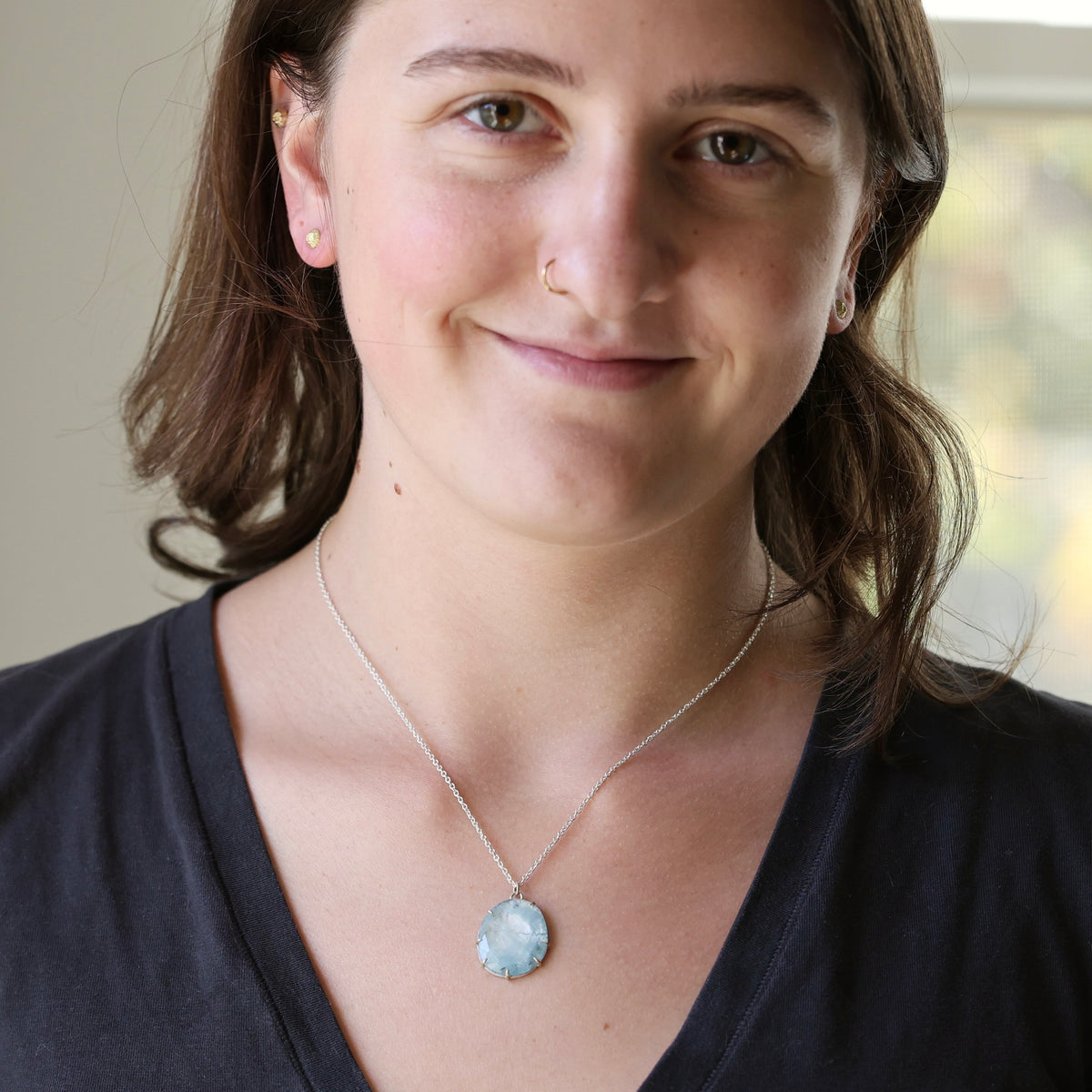 A woman wearing a silver chain necklace with a light blue oval pendant, shown in a close-up portrait.