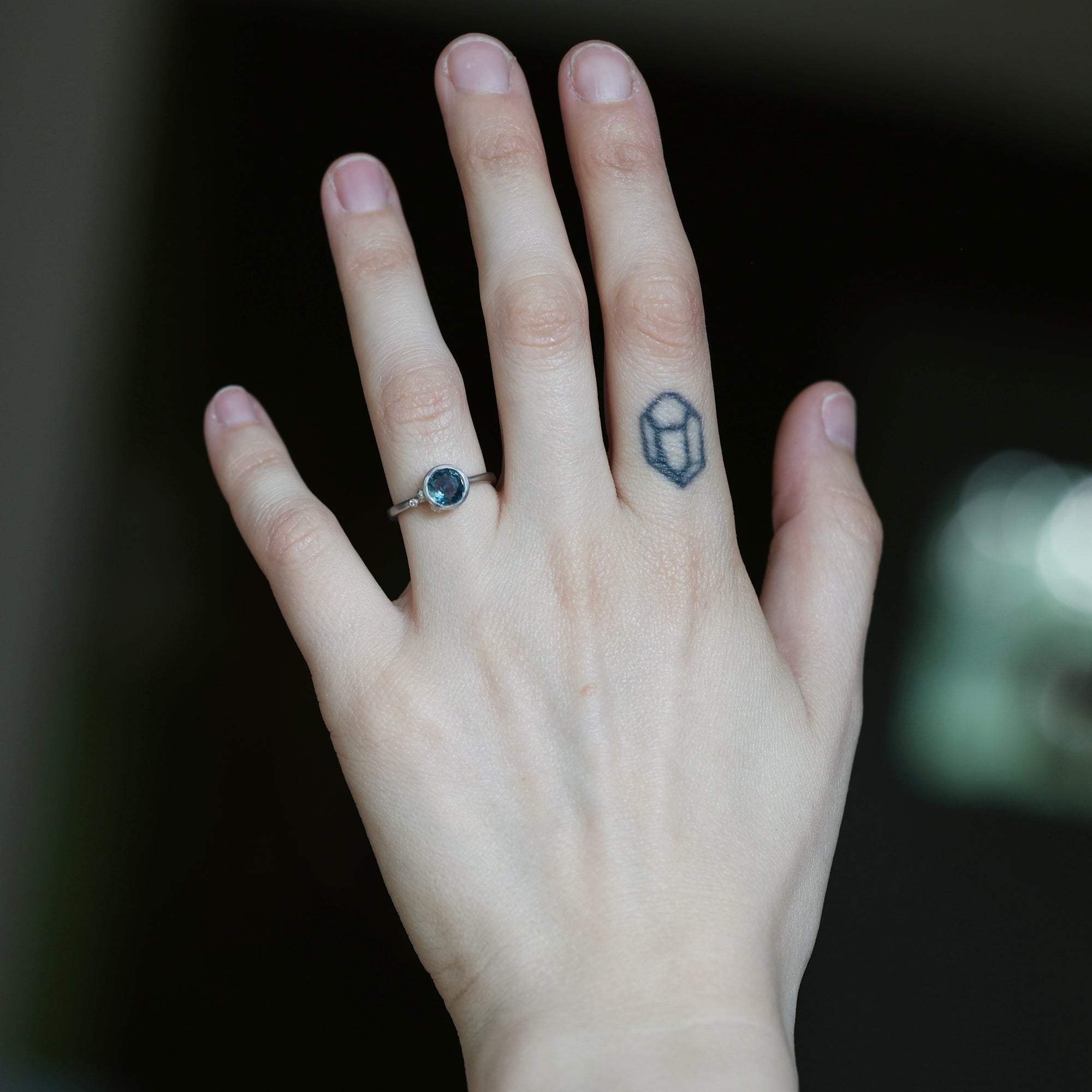A close-up of a person's hand showing a silver ring with a dark oval gemstone on the ring finger, alongside a small geometric tattoo on the middle finger.