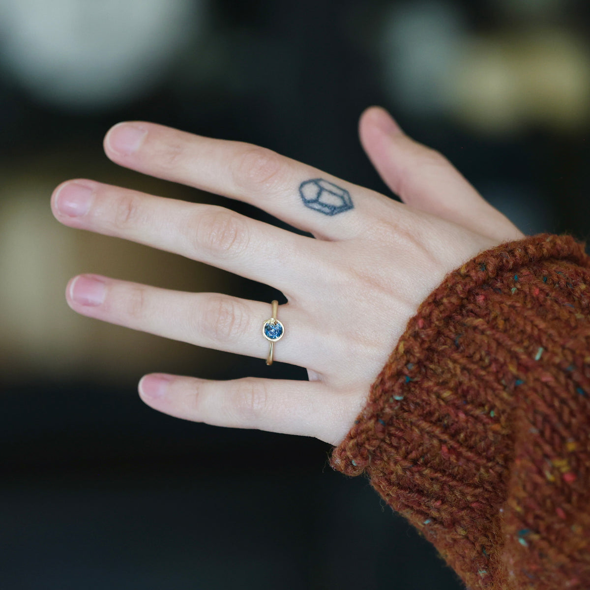 A close-up of a person's hand wearing a delicate gold ring with a small round blue gemstone, set against a dark blurred background and a brown knit sweater cuff.