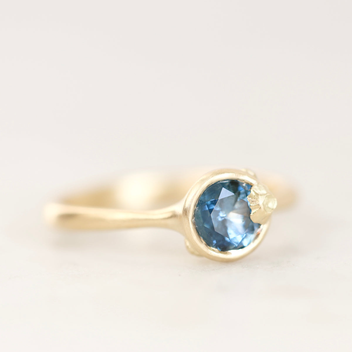 A gold ring featuring a round faceted light blue gemstone in a bezel setting, photographed on a soft white background.