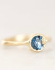 A gold ring featuring a round faceted light blue gemstone in a bezel setting, photographed on a soft white background.