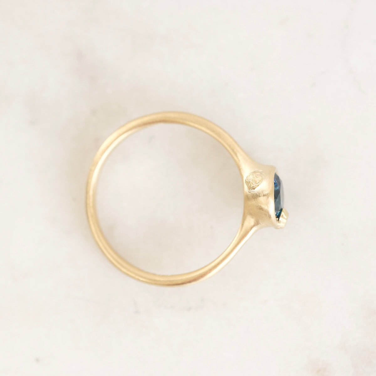 A gold ring with a small oval blue gemstone, photographed from above on a light marble surface.