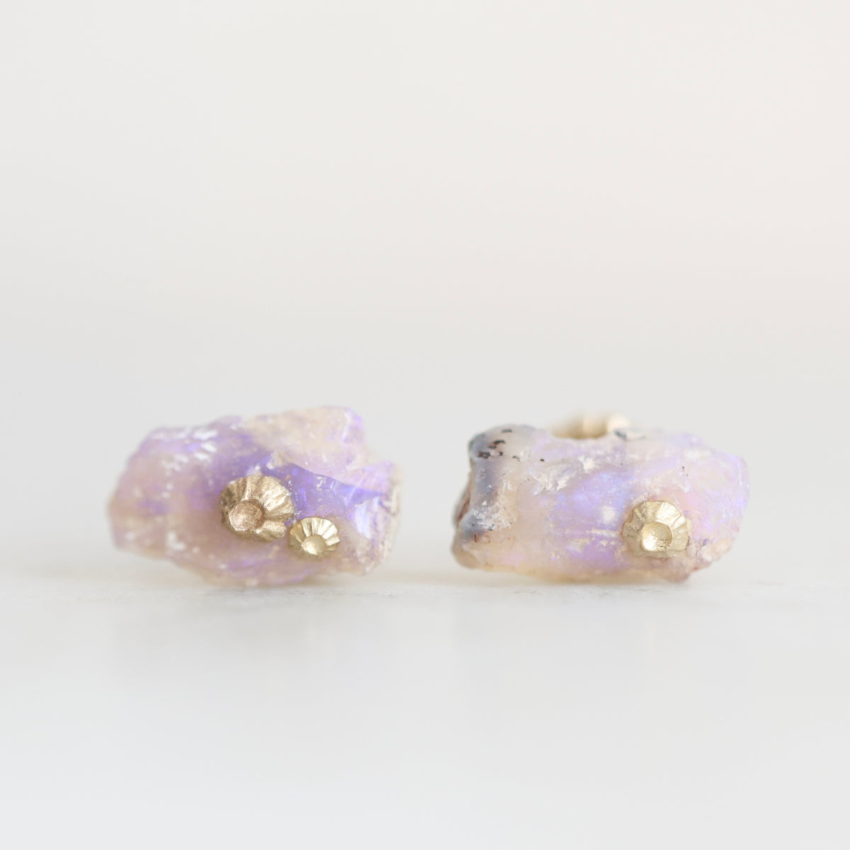 A pair of stud earrings featuring raw, iridescent opal pieces with a small gold barnacle accent on each, set against a plain white background.
