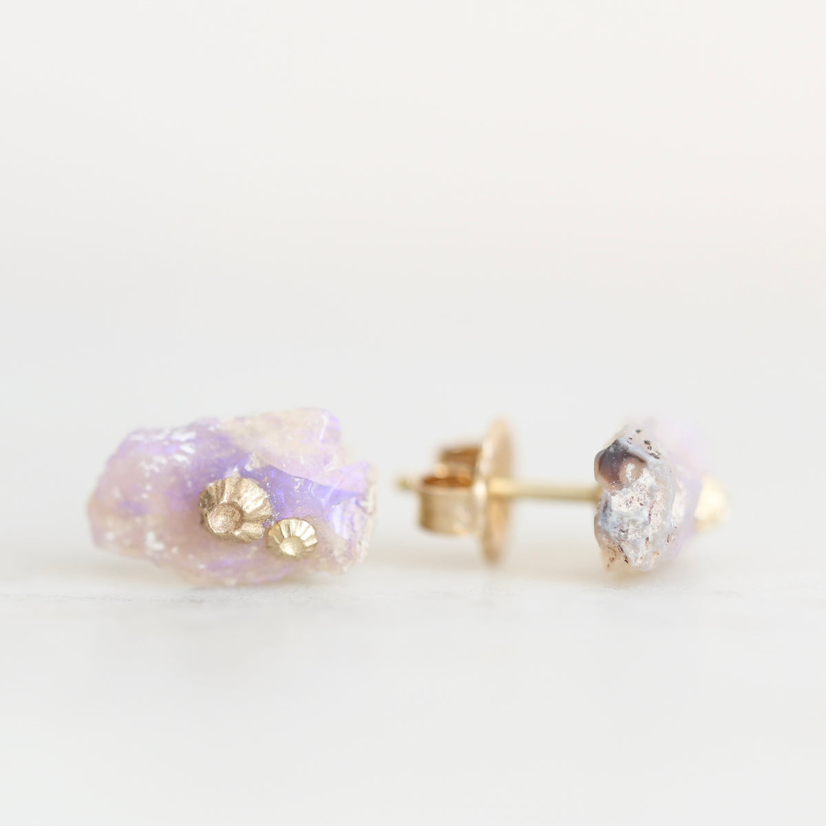 A pair of gold stud earrings featuring raw, iridescent lavender-purple opals with small gold foil accents, shown on a clean white background.