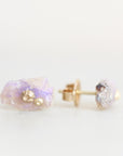 A pair of gold stud earrings featuring raw, iridescent lavender-purple opals with small gold foil accents, shown on a clean white background.