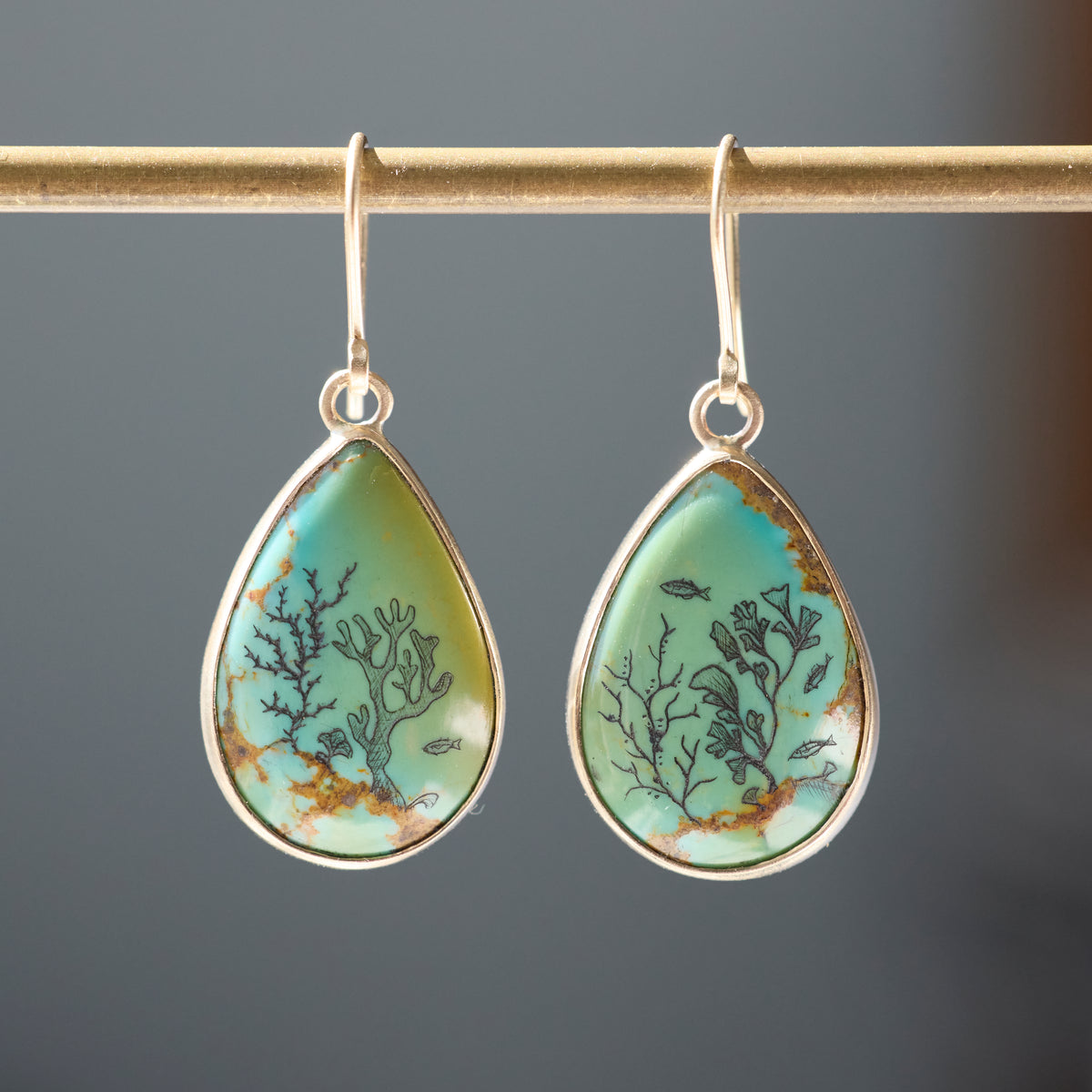 A pair of teardrop-shaped drop earrings with turquoise-blue and green stone inlays featuring black coral and fish motifs, set in silver-tone metal hoops, hanging from a gold-toned rod against a dark gray background.