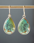 A pair of teardrop-shaped drop earrings with turquoise-blue and green stone inlays featuring black coral and fish motifs, set in silver-tone metal hoops, hanging from a gold-toned rod against a dark gray background.