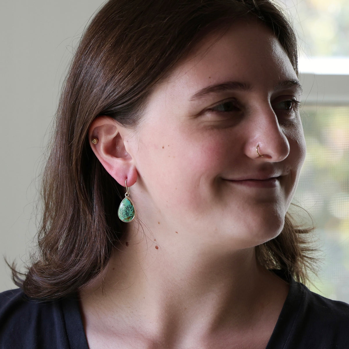 A woman with brown hair is shown in profile wearing a small gold hoop earring and a dangling teardrop-shaped green stone earring.