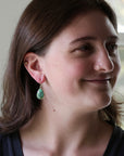 A woman with brown hair is shown in profile wearing a small gold hoop earring and a dangling teardrop-shaped green stone earring.