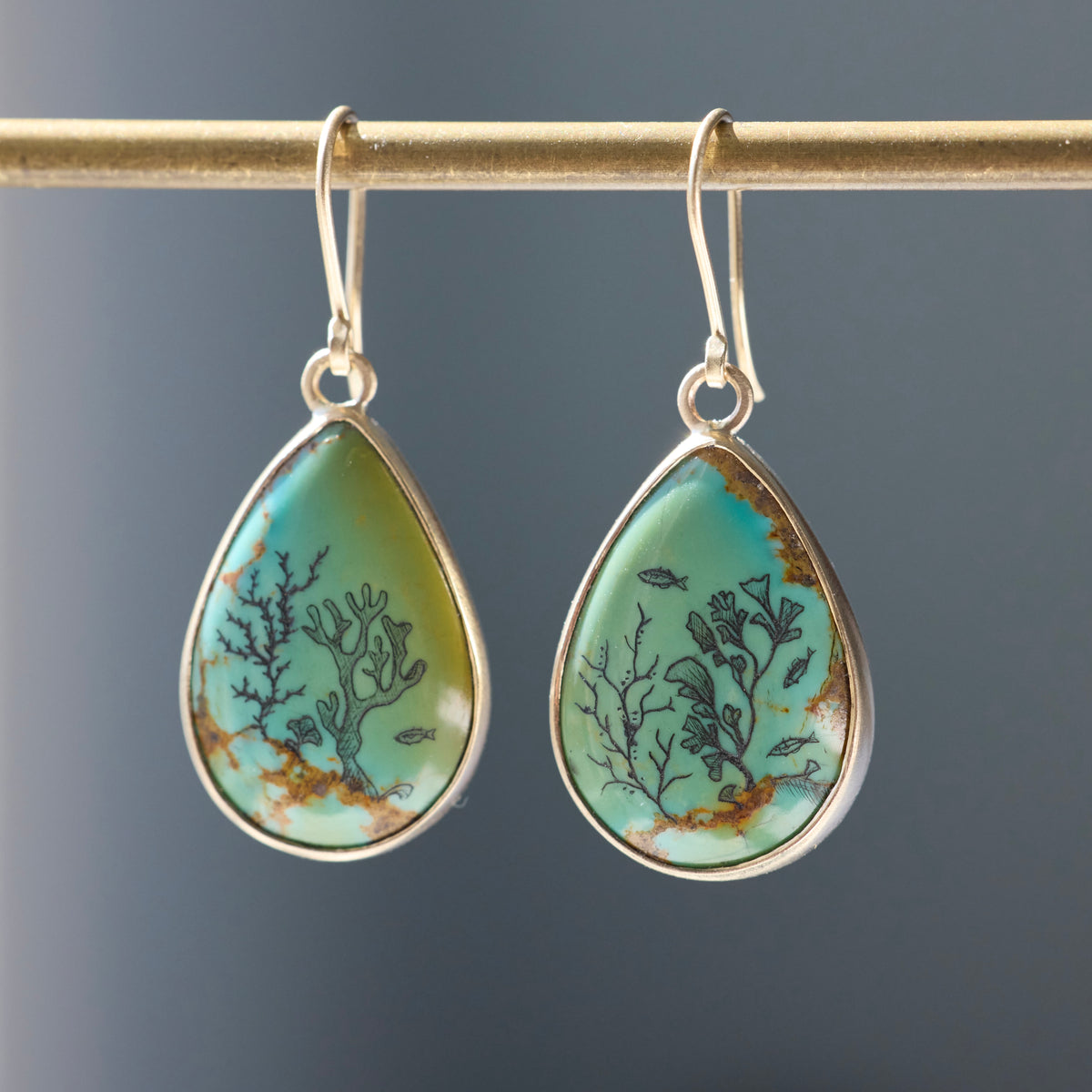 A pair of teardrop-shaped dangle earrings hanging from a gold rod. The earrings have silver-toned metal settings and hooks, featuring a light turquoise and green stone inlay with black coral and fish motifs and subtle brown veining, set against a dark gray background.