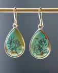 A pair of teardrop-shaped dangle earrings hanging from a gold rod. The earrings have silver-toned metal settings and hooks, featuring a light turquoise and green stone inlay with black coral and fish motifs and subtle brown veining, set against a dark gray background.