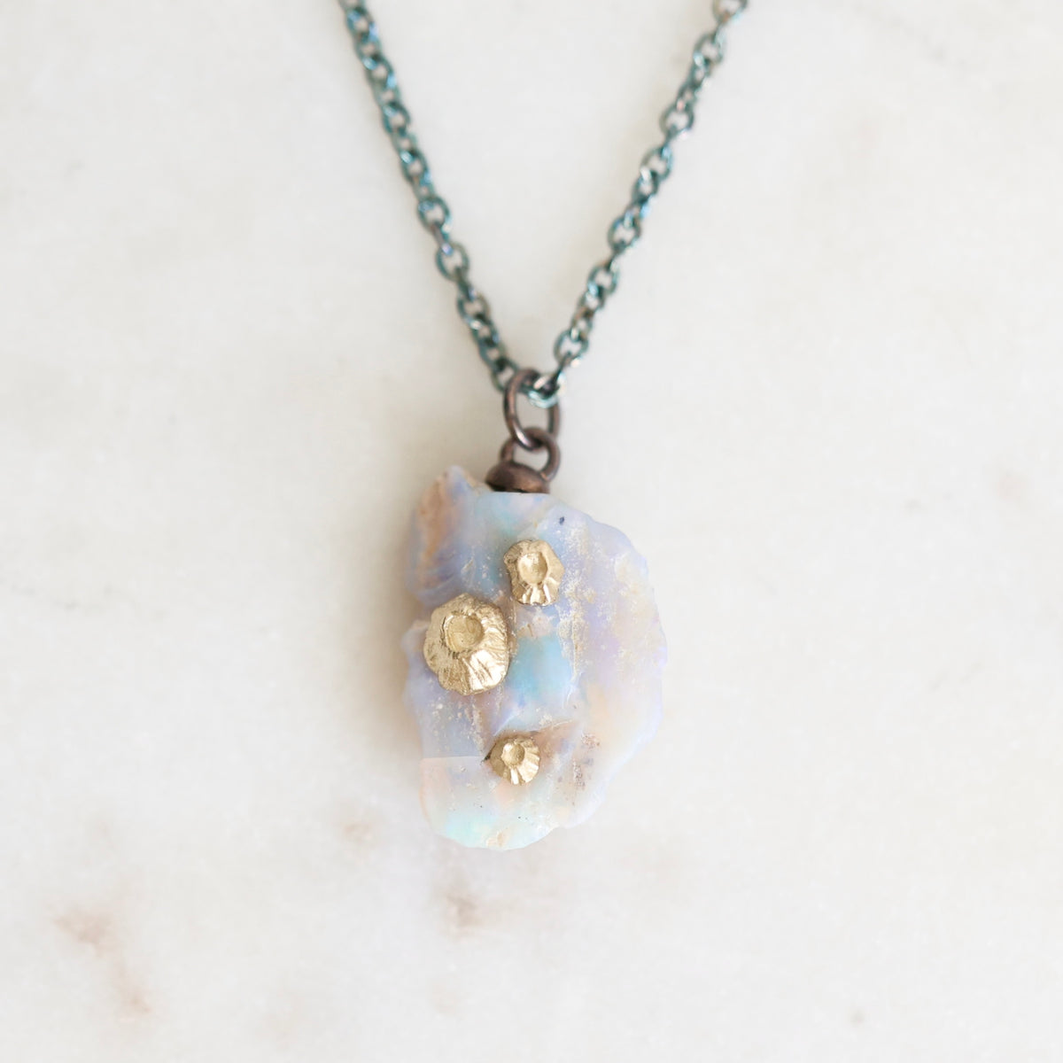 Fossilized opal pendant with gold barnacle details on an oxidized silver chain. Laying against marble background. 