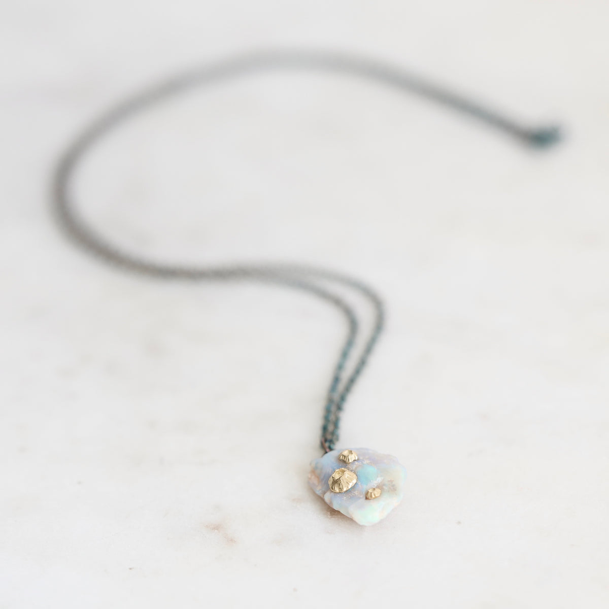 Fossilized opal pendant with gold barnacle details on an oxidized silver chain. Laying against marble background. 