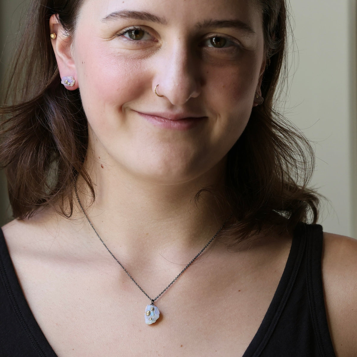 Woman wearing raw opal necklace. She has short brown hair and is standing inside smiling at the camera. 