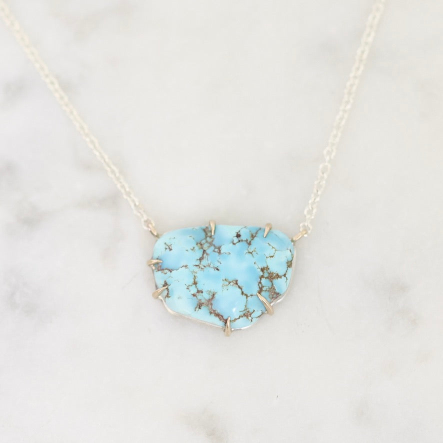 A light blue turquoise stone pendant necklace with a gold-toned chain, displayed on a white marble surface.