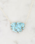 A light blue turquoise stone pendant necklace with a gold-toned chain, displayed on a white marble surface.