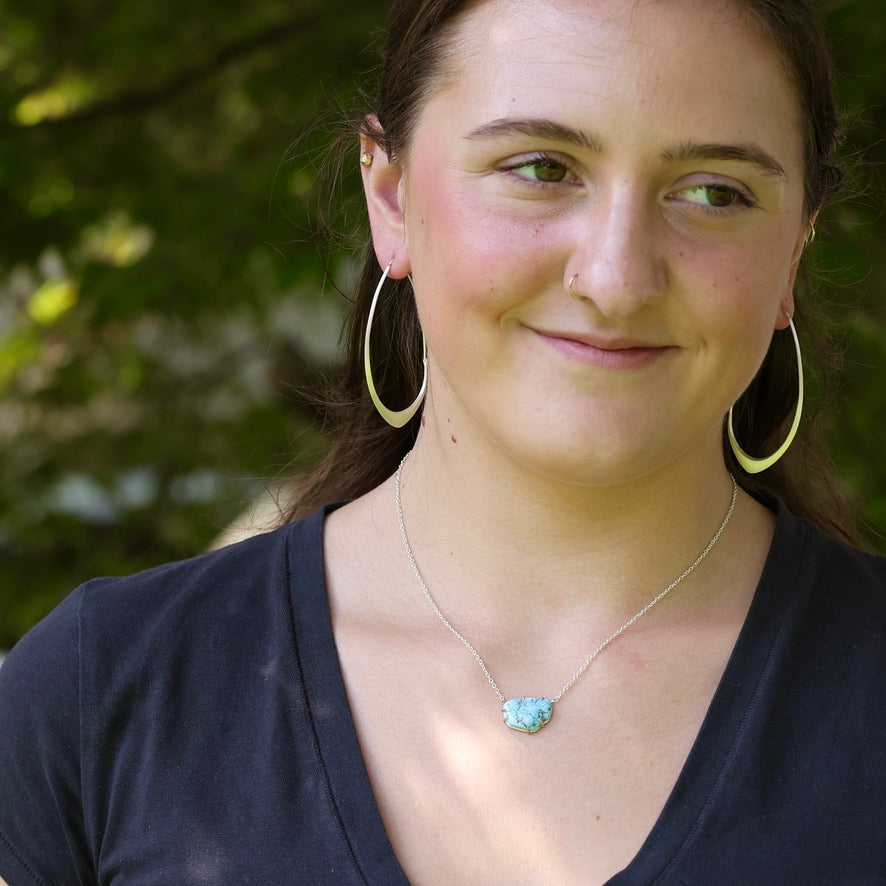 A woman wearing a silver necklace with a light blue stone pendant, gold hoop earrings, and a nose ring, photographed outdoors against a blurred green background.