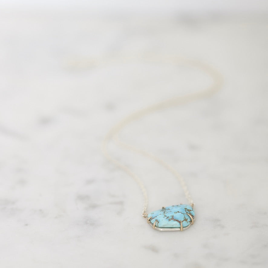 A delicate gold chain necklace featuring a small, organic-shaped turquoise stone pendant with gold tree-like branch details, resting on a light marble surface.