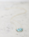 A delicate gold chain necklace featuring a small, organic-shaped turquoise stone pendant with gold tree-like branch details, resting on a light marble surface.
