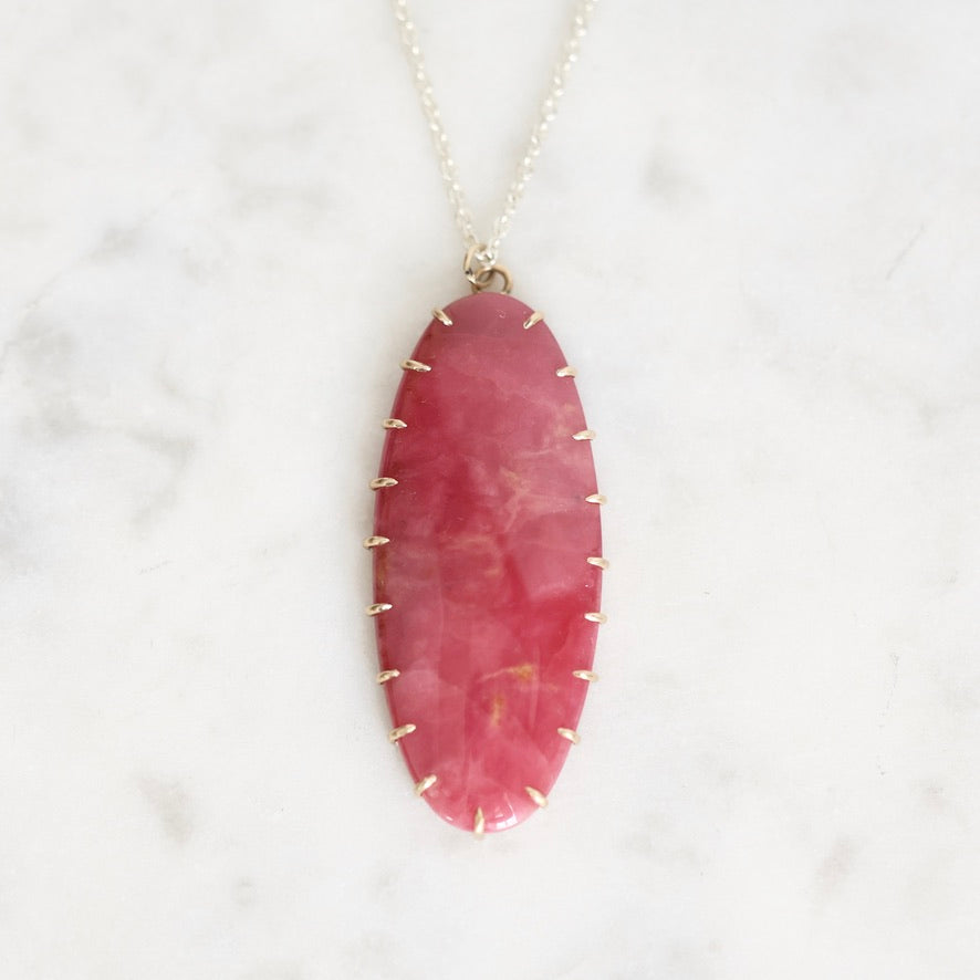 An oval pink gemstone pendant necklace with a gold-toned bezel and chain, displayed on a white marble surface.