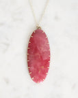 An oval pink gemstone pendant necklace with a gold-toned bezel and chain, displayed on a white marble surface.