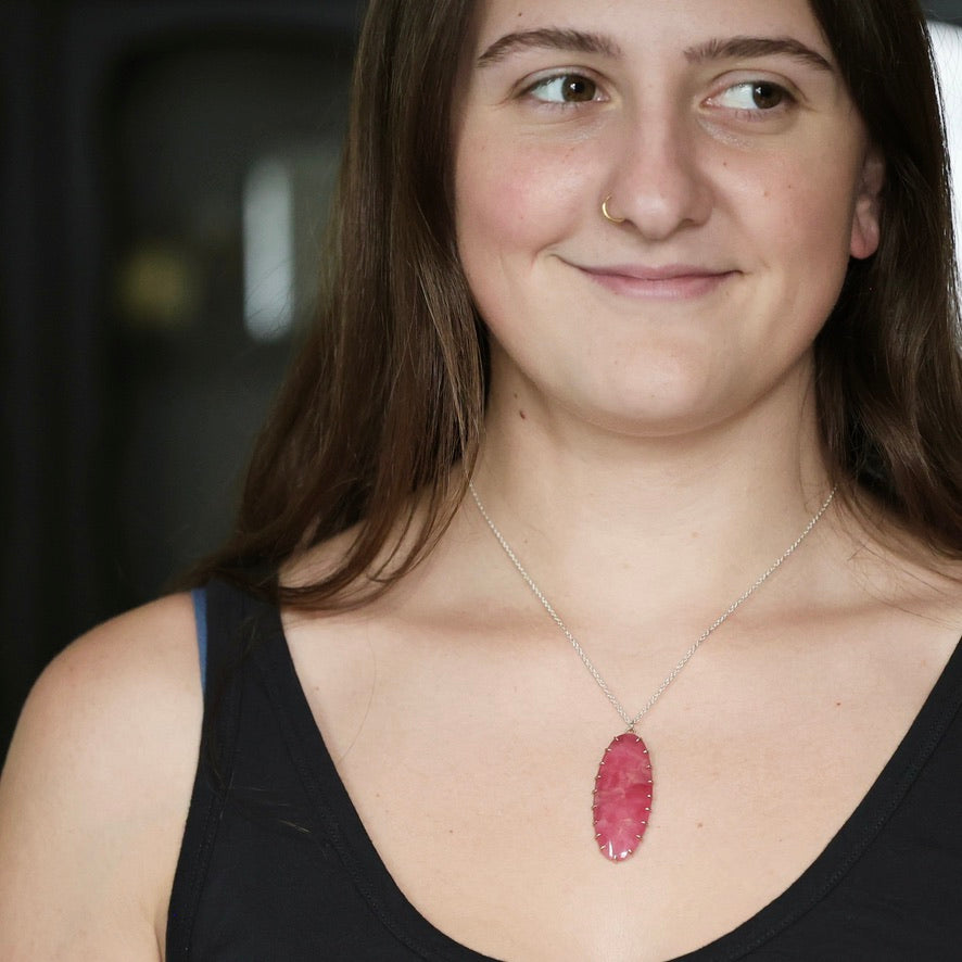 A woman wearing a silver chain necklace with a polished pink oval stone pendant, shown in a close-up portrait.