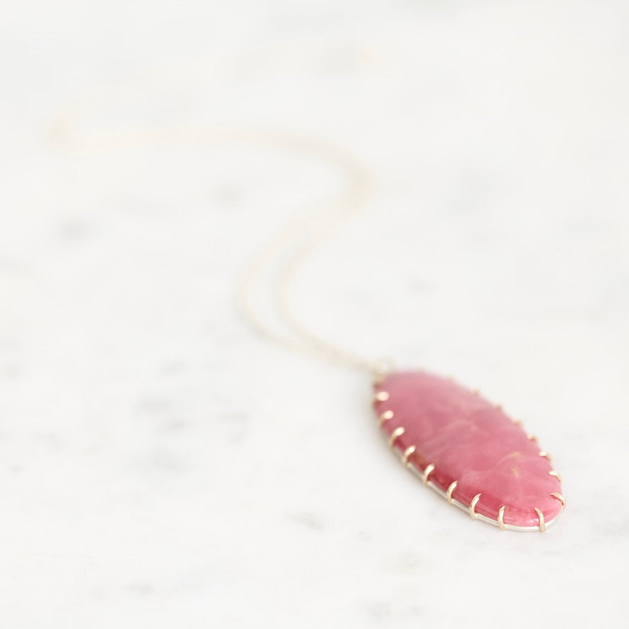 A delicate gold chain necklace with an oval pink stone pendant, resting on a light marble surface.