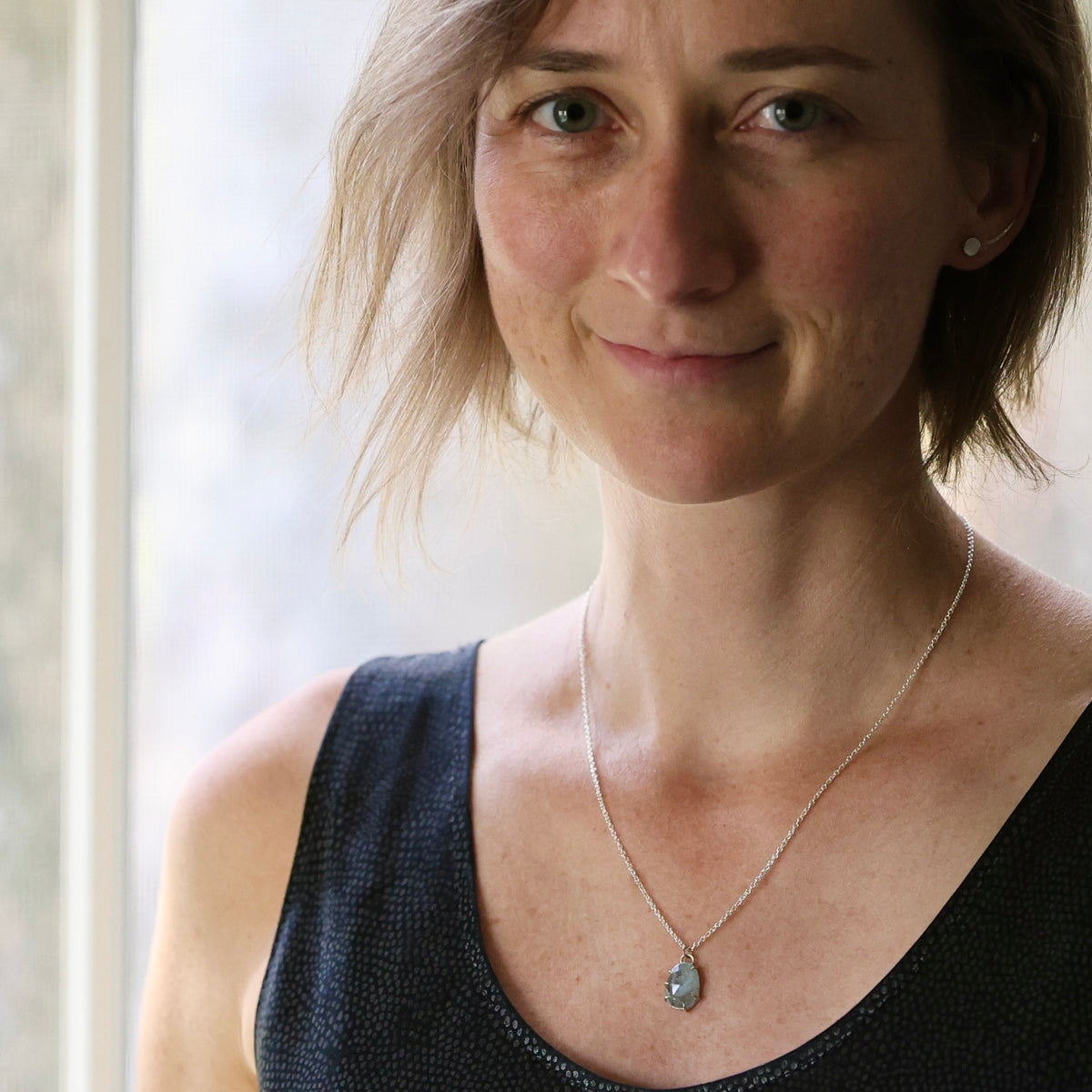 A woman with short blonde hair wearing a delicate silver necklace with a teardrop-shaped gray stone pendant, photographed in soft natural light near a window.