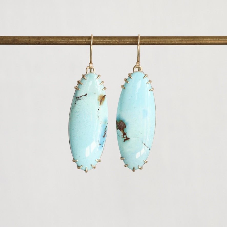 A pair of drop earrings featuring oval light blue turquoise stones with natural brown matrix, suspended from gold hooks and set in gold-toned metal prong settings, hanging from a gold-colored horizontal bar against a light gray background.