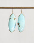 A pair of drop earrings featuring oval light blue turquoise stones with natural brown matrix, suspended from gold hooks and set in gold-toned metal prong settings, hanging from a gold-colored horizontal bar against a light gray background.