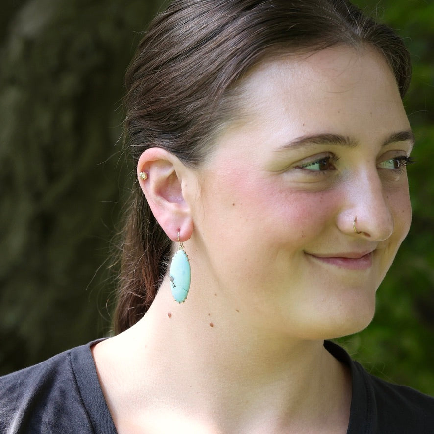 A woman wearing a light blue-green teardrop-shaped dangle earring with a gold-toned wire loop, photographed outdoors against a blurred green background.