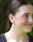 A woman wearing a light blue-green teardrop-shaped dangle earring with a gold-toned wire loop, photographed outdoors against a blurred green background.