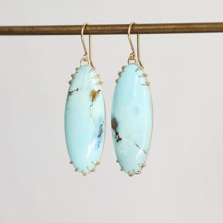 A pair of gold drop earrings with oval light blue turquoise-like stones, hanging from a horizontal gold rod against a plain light gray background.