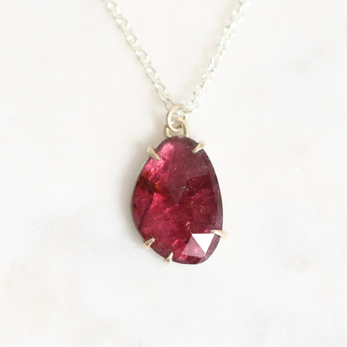 A teardrop-shaped faceted dark red gemstone pendant in a silver bezel setting, hanging from a delicate silver chain against a light off-white marble background.