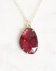 A teardrop-shaped faceted dark red gemstone pendant in a silver bezel setting, hanging from a delicate silver chain against a light off-white marble background.