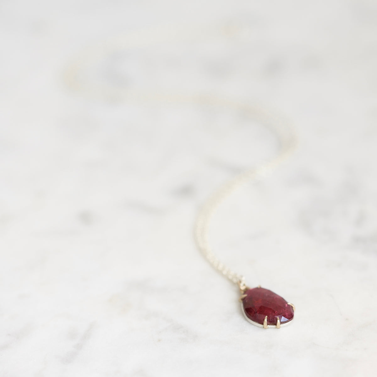 A delicate gold chain necklace with a small, teardrop-shaped deep red gemstone pendant, resting on a light marble surface.