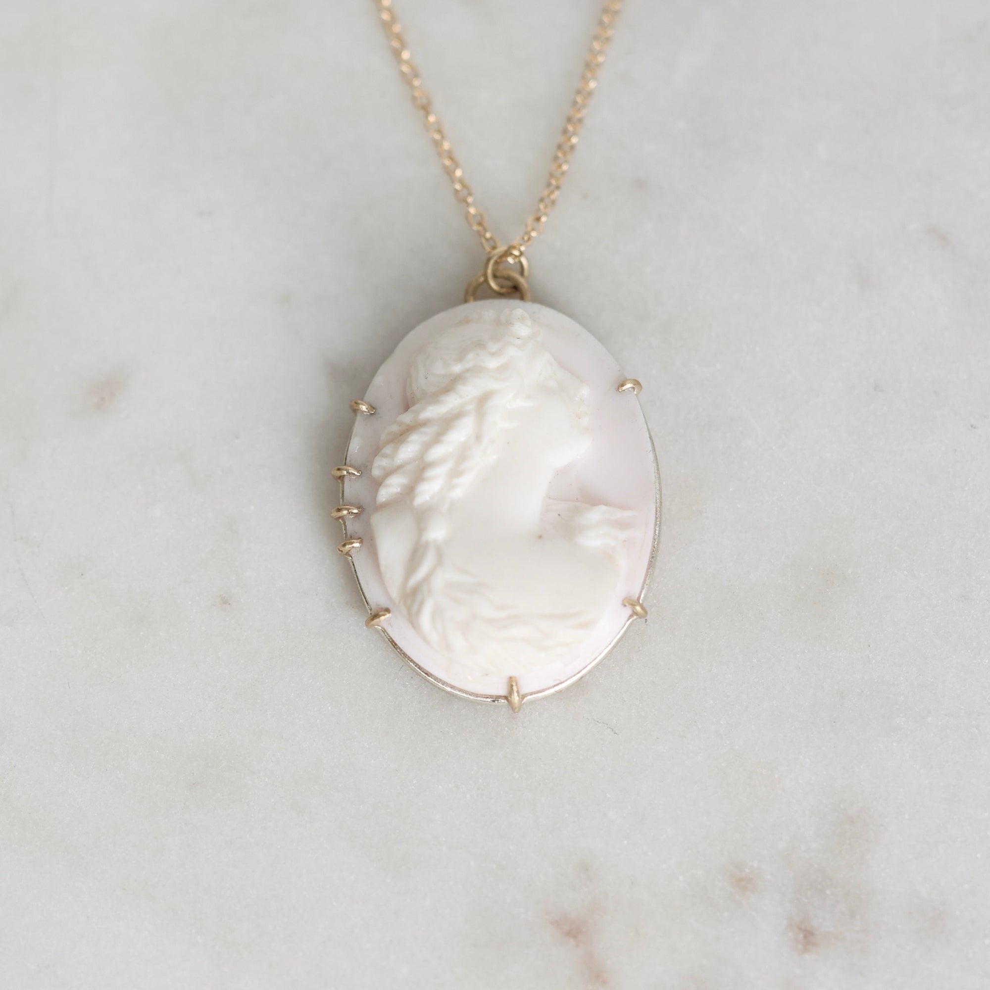 A gold chain necklace with an oval carved cameo pendant, displayed on a light marble surface. The pendant features a profile of a woman in cream and white with gold prong settings.
