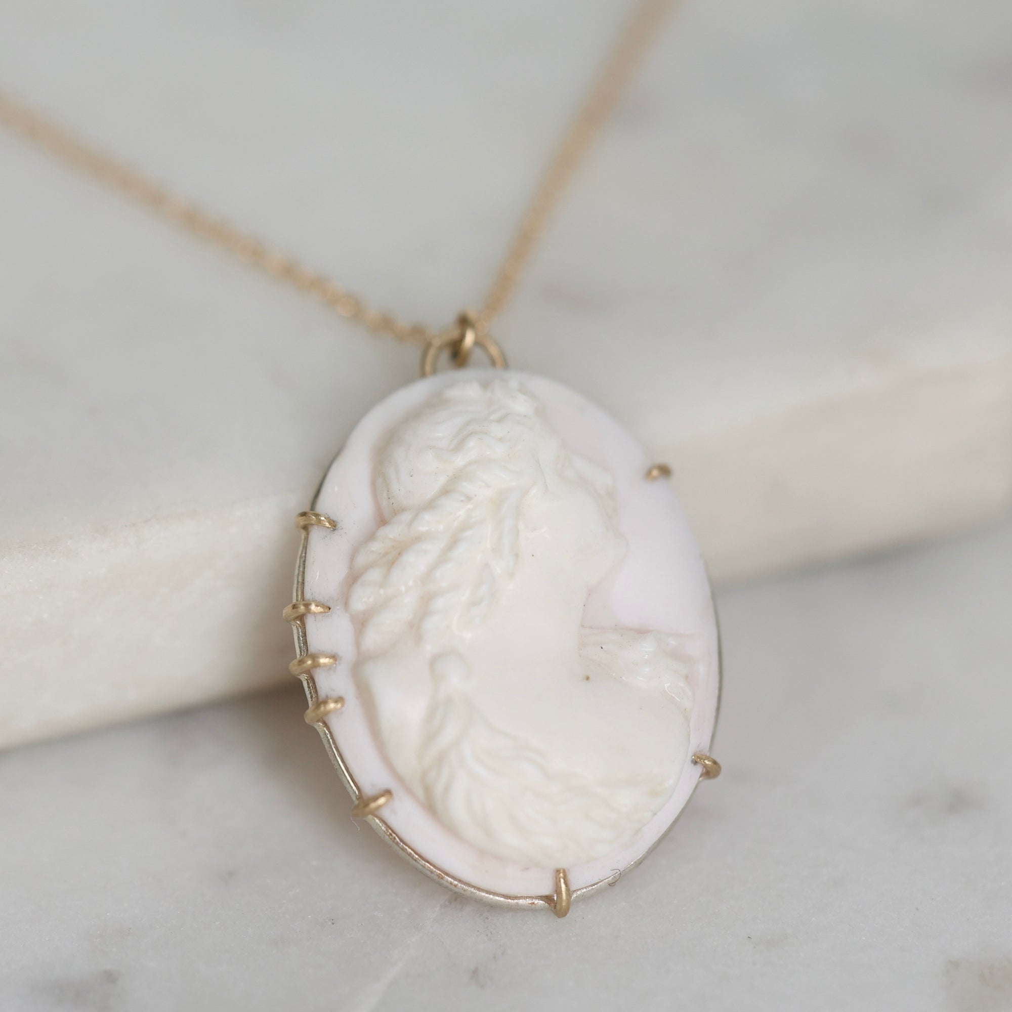 A gold-tone necklace with an oval pendant featuring a white carved cameo-style portrait, resting on a light marble surface.
