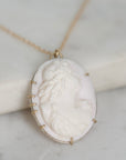 A gold-tone necklace with an oval pendant featuring a white carved cameo-style portrait, resting on a light marble surface.