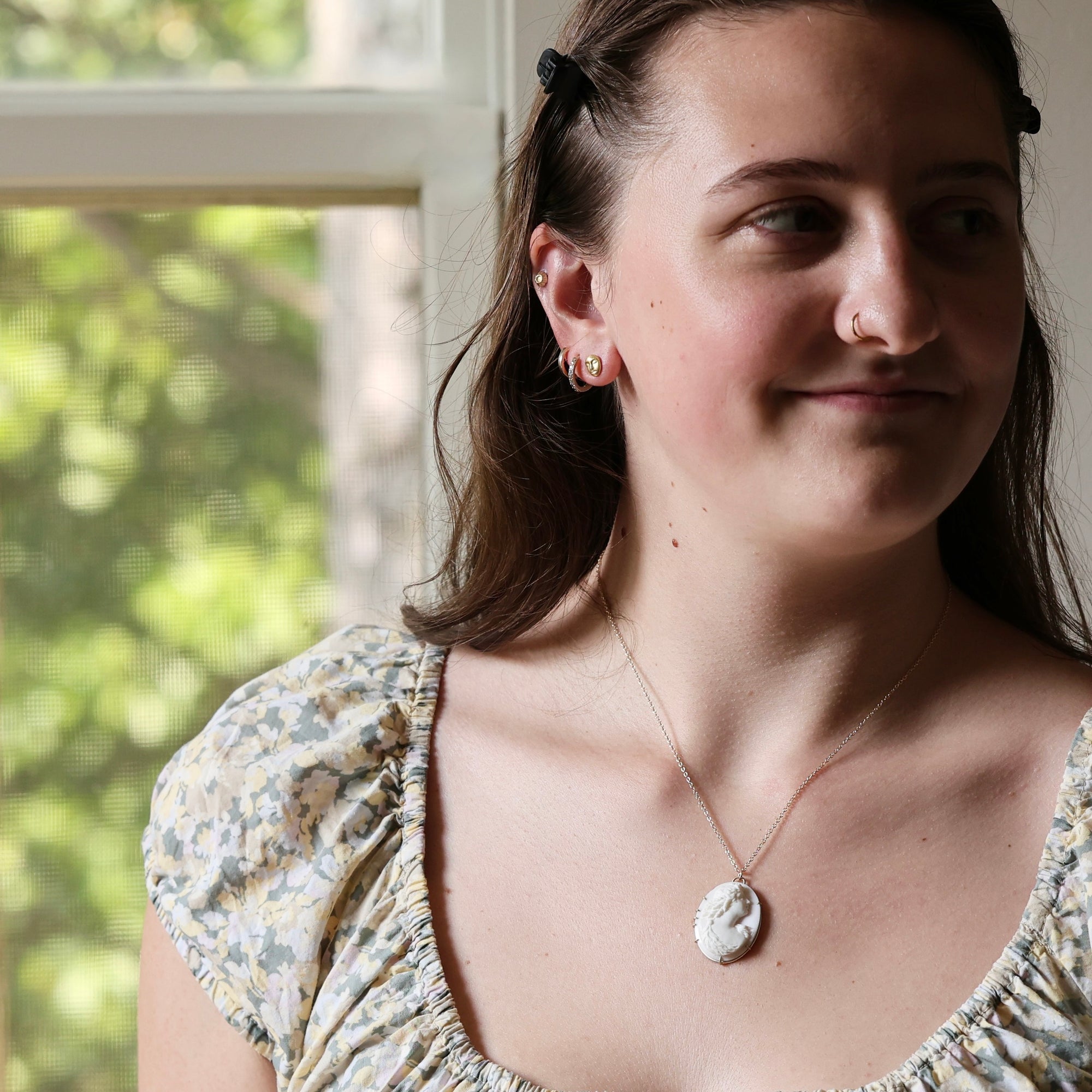 A woman wearing a delicate gold chain necklace with a white shell pendant, photographed in a bright indoor setting with a window in the background.