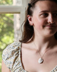 A woman wearing a delicate gold chain necklace with a white shell pendant, photographed in a bright indoor setting with a window in the background.
