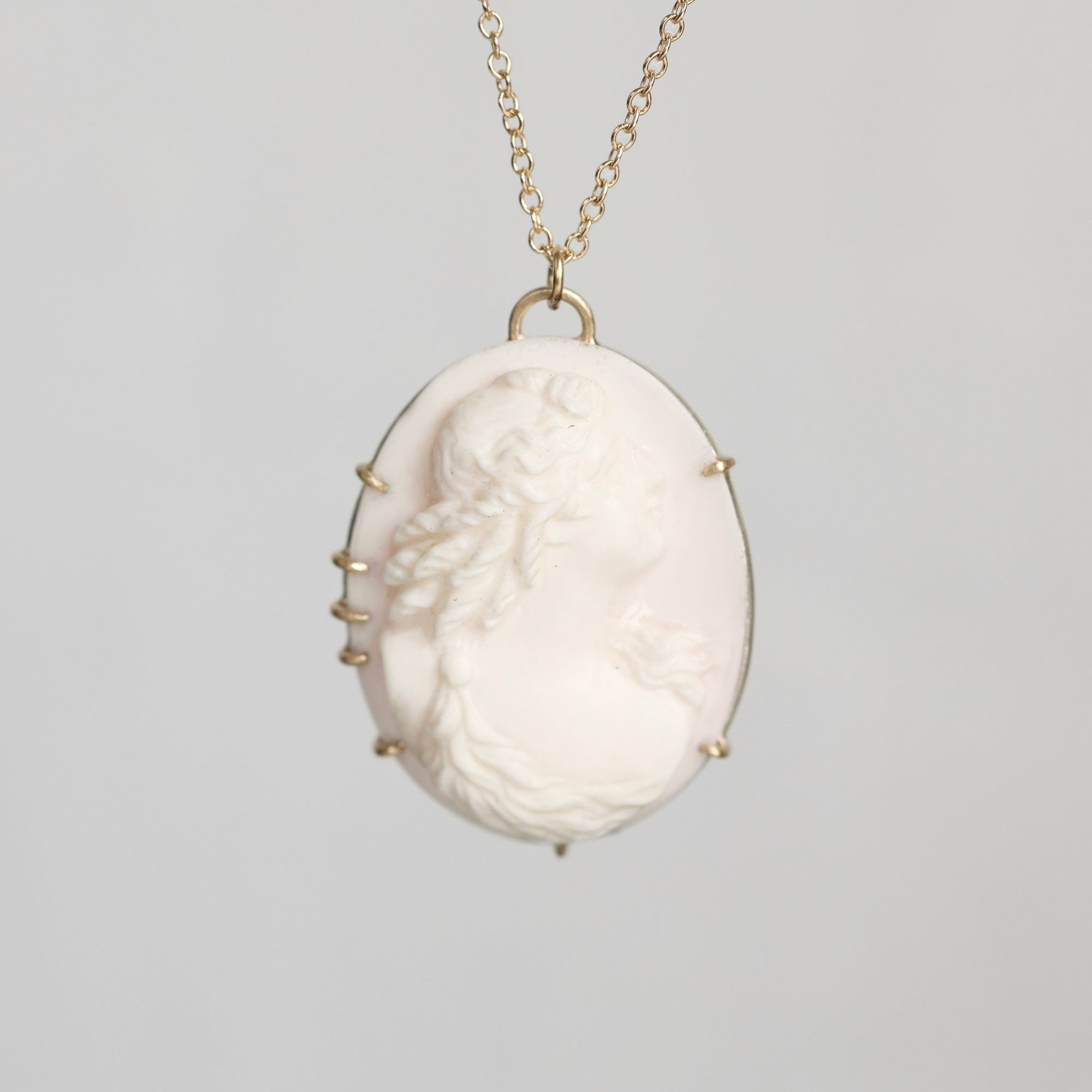 A gold chain necklace featuring an oval white carved cameo pendant with a classical profile design, set in a gold bezel with small decorative side prongs, shown against a neutral light gray background.