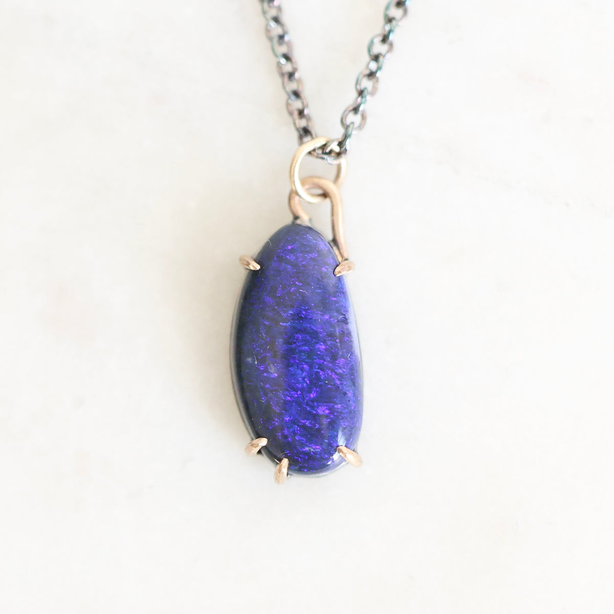 Dark colored irregularly shaped opal set in gold prongs against a plain background.